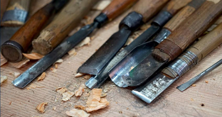 Eine Auwahl an Spatel und anderen Werkzeugen zum Holzbearbeiten liegen auf einem Tisch. Holzspähne lassen darauf schließen, dass sie kürzlich benutzt wurden.