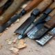 Eine Auwahl an Spatel und anderen Werkzeugen zum Holzbearbeiten liegen auf einem Tisch. Holzspähne lassen darauf schließen, dass sie kürzlich benutzt wurden.