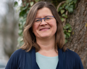 Krisztina Willy steht vor einem Efoi bewachsenen Baumstamm. Sie trägt ein pastellfarbenes Top und einen dunkelblaue Jacke darüber.